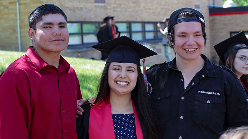 A young Native American graduate smiles broadly in her cap and gown with a friend on either side.